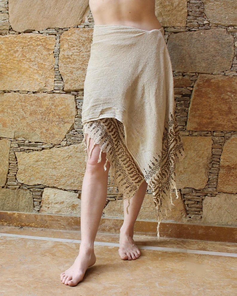 May include: A beige wrap skirt featuring a textured fabric with a dark brown printed border pattern along the asymmetrical hemline. The garment includes decorative fringe detailing at the edges and is worn wrapped around the waist.