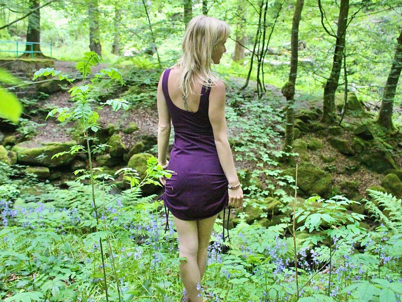 May include: A woman wearing a purple, form-fitting dress stands in a lush green forest. The dress has a low back and a cinched waist. The woman is surrounded by green foliage and blue flowers.