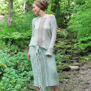 May include: A long-sleeved, light-coloured top with a brown geometric diamond pattern down the centre and on the cuffs, paired with a light green wrap skirt featuring a dark green printed vertical border and a fringed edge along the side.