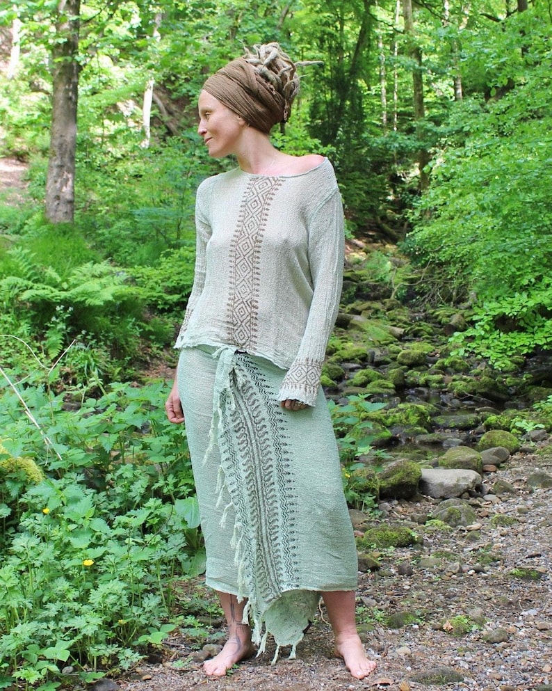 May include: A long-sleeved, light-coloured top with a brown geometric diamond pattern down the centre and on the cuffs, paired with a light green wrap skirt featuring a dark green printed vertical border and a fringed edge along the side.