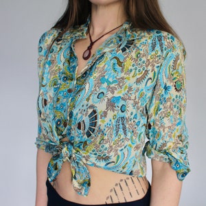 Op de afbeelding: Een blauw en wit paisley-print overhemd met een bruine bies. Het overhemd is in de taille geknoopt en heeft lange mouwen.