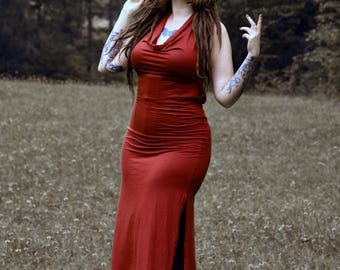 May include: A sleeveless, cowl-neck, rust-coloured midi dress with a side slit, paired with a black wide-brimmed hat featuring a white pentagram and bone-like illustrations, worn over black leggings and dark boots.