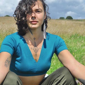 May include: A short-sleeved teal crop top with a V-neckline paired with olive green pants. The top is made of a solid-colored fabric and features a fitted silhouette that ends above the waistline, while the pants have a relaxed, loose-fitting cut.