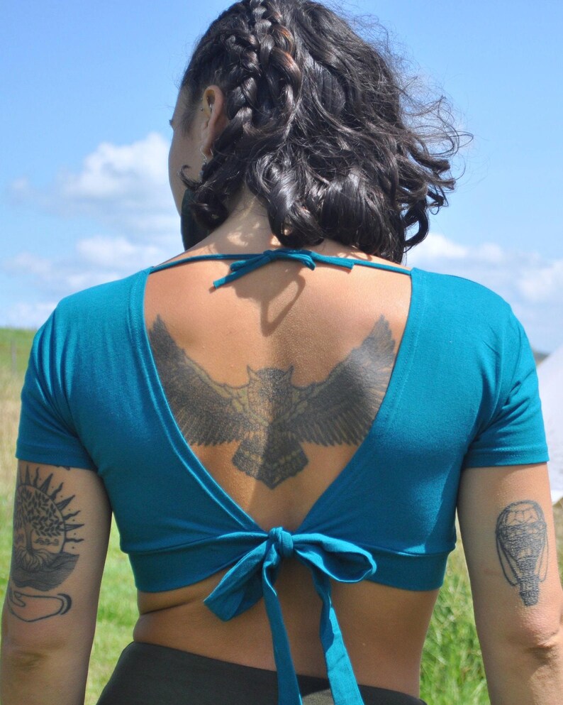 May include: A teal short-sleeved crop top featuring a deep open back design with a tie closure at the nape of the neck and a matching tie closure at the lower back, constructed from a solid-colored fabric with a fitted silhouette.