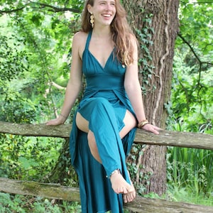 Puede incluir: Un vestido turquesa con cuello halter, escote pronunciado y falda fluida. El vestido se lleva al aire libre, con la persona sentada en una valla de madera. El fondo presenta una exuberante vegetación y un tronco de árbol.