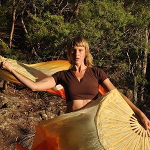 May include: A dark brown short-sleeved V-neck crop top paired with two large, flowing silk fans. The fans feature a gradient color pattern transitioning from bright yellow to vibrant orange, supported by light-colored wooden ribs.