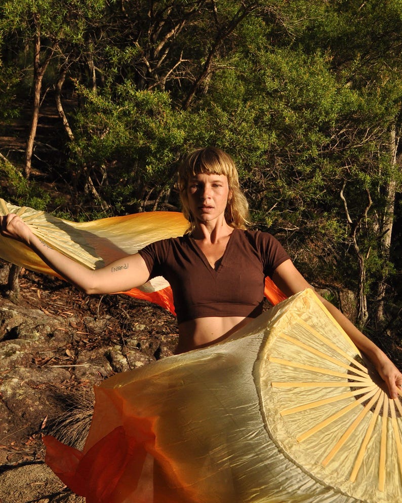 May include: A dark brown short-sleeved V-neck crop top paired with two large, flowing silk fans. The fans feature a gradient color pattern transitioning from bright yellow to vibrant orange, supported by light-colored wooden ribs.
