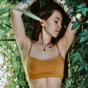 May include: A woman with tattoos on her arms is wearing a mustard yellow crop top and a white skirt. She is standing in front of a wall of green leaves.