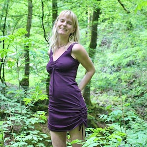 May include: A woman with blonde hair is wearing a purple, sleeveless dress with a gathered neckline. She is standing in a forest setting.