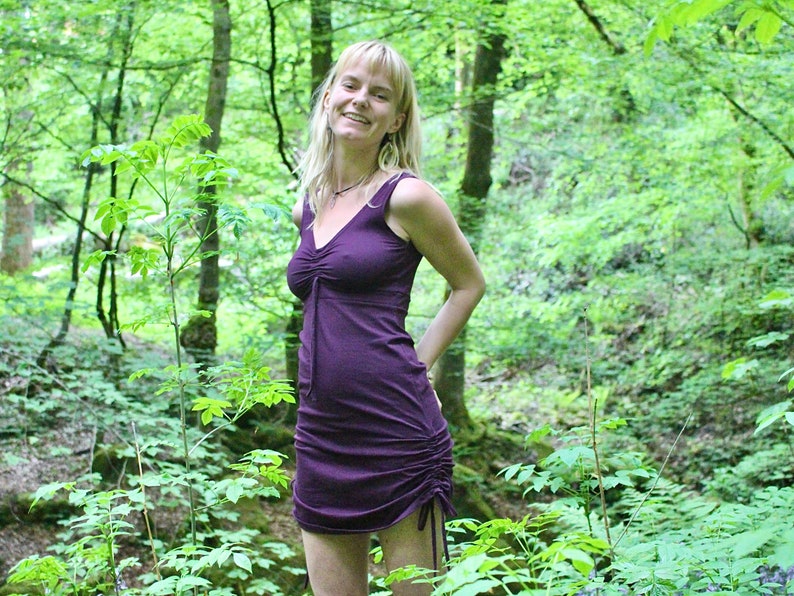 May include: A woman with blonde hair is wearing a purple, sleeveless dress with a gathered neckline. She is standing in a forest setting.