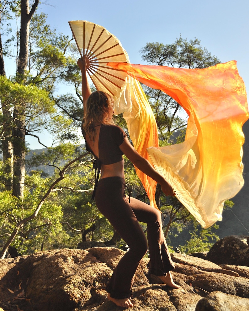 May include: A large handheld folding fan with long, flowing silk fabric in a gradient of yellow, orange, and red. The fan features a wooden frame with multiple ribs that extend to support the lightweight, translucent material.