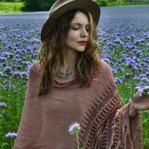 May include: A dusty rose knit poncho featuring a dark brown geometric pattern along the hem and fringe detailing. The garment has a loose cowl neckline and a textured weave. A wide-brimmed tan felt hat is worn above the shoulders.