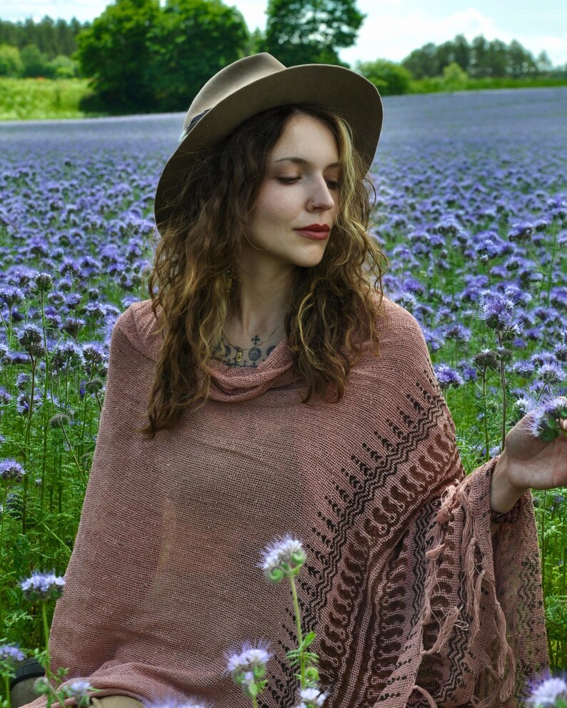 May include: A dusty rose knit poncho featuring a dark brown geometric pattern along the hem and fringe detailing. The garment has a loose cowl neckline and a textured weave. A wide-brimmed tan felt hat is worn above the shoulders.