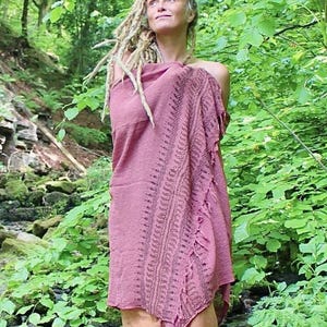 May include: A dusty rose-coloured wrap or shawl featuring a textured fabric with a dark, intricate, repeating geometric border pattern along one side and a frayed, fringed edge, draped over the shoulders and upper body in a loose, flowing style.