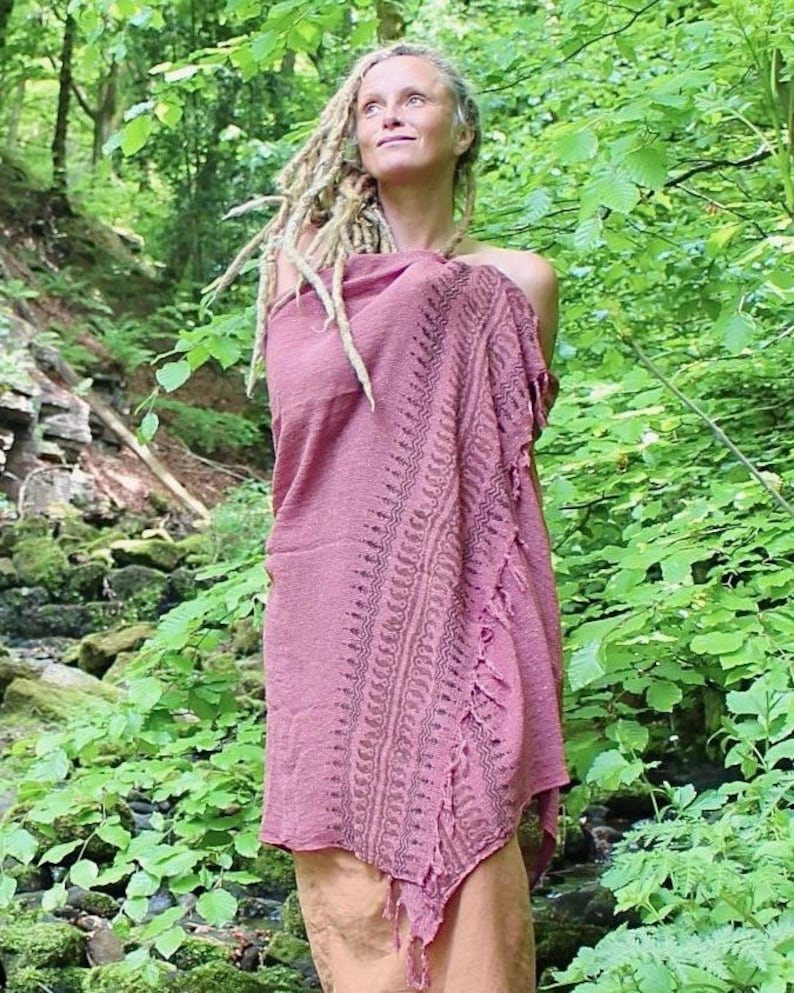May include: A dusty rose-coloured wrap or shawl featuring a textured fabric with a dark, intricate, repeating geometric border pattern along one side and a frayed, fringed edge, draped over the shoulders and upper body in a loose, flowing style.