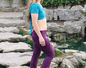 May include: A teal short-sleeved wrap top with a tie closure and deep purple flared pants featuring a ruffled hem. The pants have a drawstring waist and a fitted silhouette through the hips and thighs, transitioning into a wide flare at the bottom.