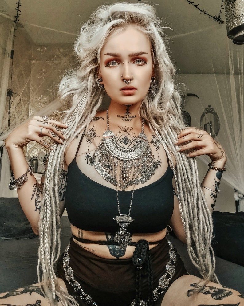 May include: A woman with long, white dreadlocks wears a black vest top and a silver, layered necklace with a large pendant. She has multiple piercings and tattoos, including a nose ring and a septum ring.