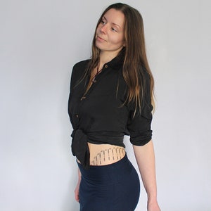 Puede incluir: Una mujer con una camisa negra de botones atada a la cintura y una falda azul oscuro. Tiene un peque&ntilde;o tatuaje en la parte baja del abdomen.