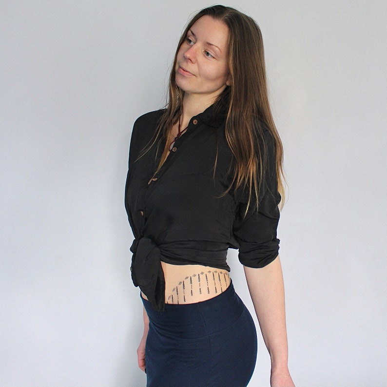 Puede incluir: Una mujer con una camisa negra de botones atada a la cintura y una falda azul oscuro. Tiene un peque&ntilde;o tatuaje en la parte baja del abdomen.