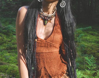 May include: A woman with long black hair wearing a brown halter top with a layered gemstone necklace and silver earrings. She is standing in a forest setting.