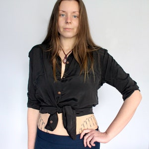Puede incluir: Una mujer con una camisa negra de botones atada a la cintura, un collar oscuro con un colgante de piedra y una falda azul oscuro. Tiene el pelo largo y casta&ntilde;o y lleva un anillo peque&ntilde;o en el dedo.