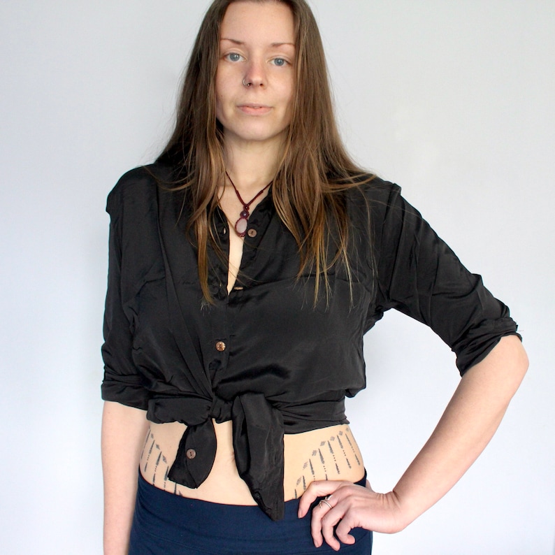 Puede incluir: Una mujer con una camisa negra de botones atada a la cintura, un collar oscuro con un colgante de piedra y una falda azul oscuro. Tiene el pelo largo y casta&ntilde;o y lleva un anillo peque&ntilde;o en el dedo.