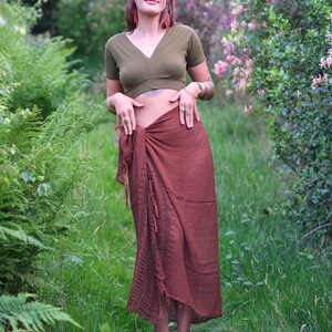 May include: An olive green short-sleeved V-neck crop top paired with a long, dark brown wrap skirt. The skirt features a textured fabric with a dark, intricate geometric pattern running vertically along the front side and frayed fringe edges.
