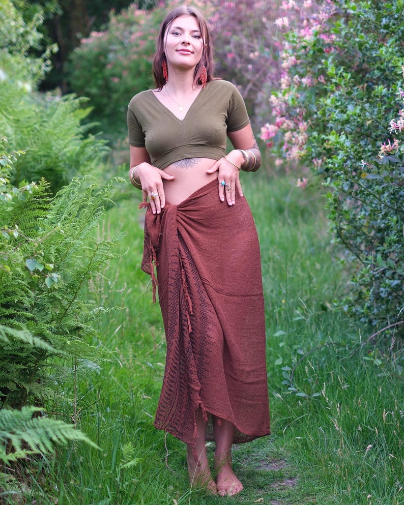 May include: An olive green short-sleeved V-neck crop top paired with a long, dark brown wrap skirt. The skirt features a textured fabric with a dark, intricate geometric pattern running vertically along the front side and frayed fringe edges.