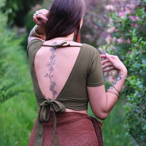 May include: An olive green short-sleeved crop top featuring a deep open back design with a thin tie closure at the neck and a wider tie closure at the waist, constructed from a solid fabric with a smooth texture and a fitted silhouette.