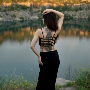 May include: A woman with long dark hair wearing a black crop top and a long black skirt. She has a large intricate tattoo on her back. She is standing by a body of water with a rocky shore and green trees in the background.
