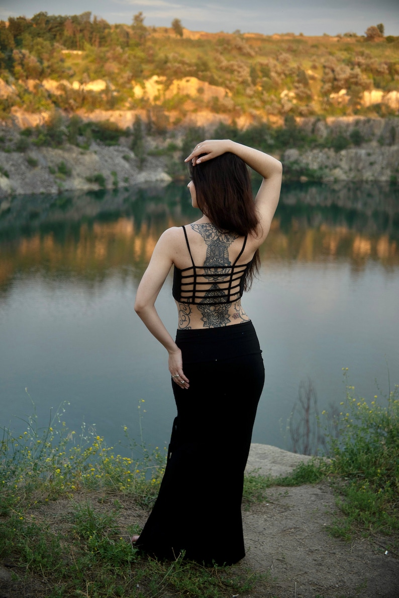 May include: A woman with long dark hair wearing a black crop top and a long black skirt. She has a large intricate tattoo on her back. She is standing by a body of water with a rocky shore and green trees in the background.