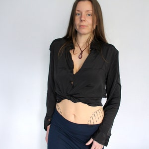 Puede incluir: Una mujer con una camisa negra de manga larga anudada a la cintura, una falda azul oscuro y un collar con un colgante. Tiene un tatuaje en la parte baja del abdomen.