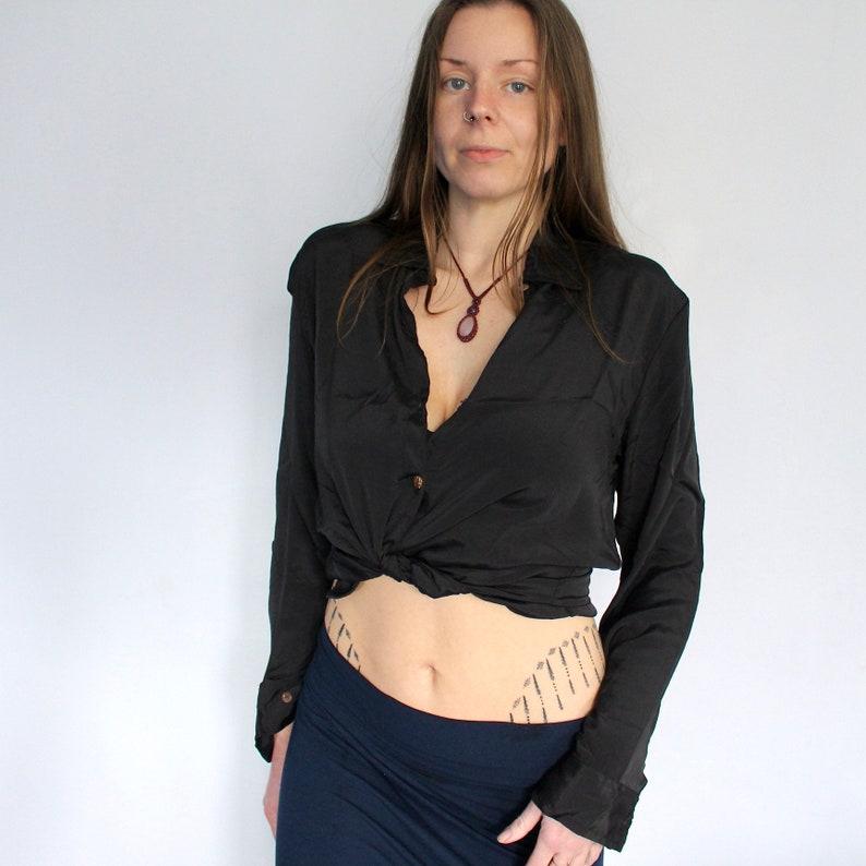 Puede incluir: Una mujer con una camisa negra de manga larga anudada a la cintura, una falda azul oscuro y un collar con un colgante. Tiene un tatuaje en la parte baja del abdomen.