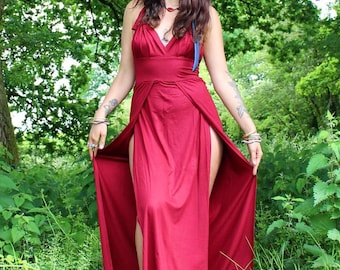 May include: A floor-length deep red dress featuring a V-neckline, a fitted bodice, and a skirt with two high side slits that reveal the legs. The garment is sleeveless with wide straps that tie at the shoulders, creating a draped effect.