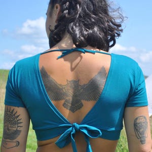 May include: A teal short-sleeved crop top featuring a deep open back design with a tie closure at the nape of the neck and a matching tie closure at the lower back, constructed from a solid-colored fabric with a fitted silhouette.
