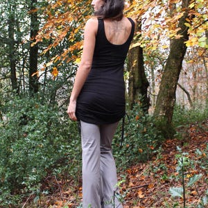 May include: A black tank top with a gathered hem and a gray pair of pants. The person is standing in a wooded area with fall foliage.