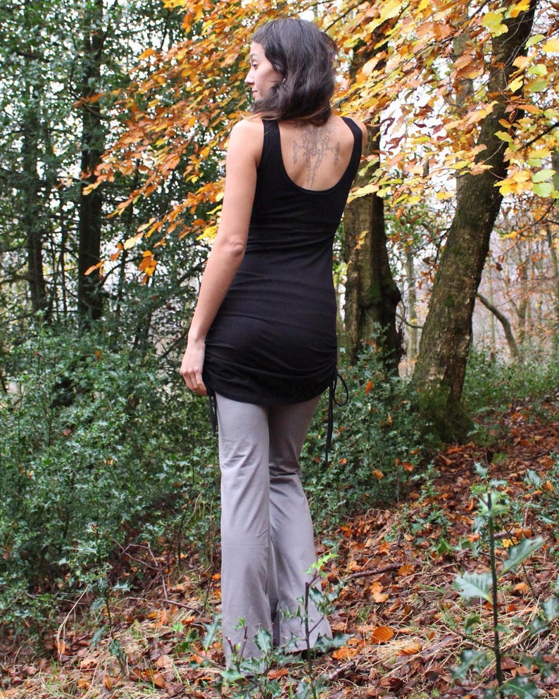 May include: A black tank top with a gathered hem and a gray pair of pants. The person is standing in a wooded area with fall foliage.