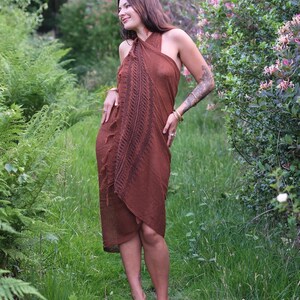 May include: A rust-coloured wrap dress featuring a halter neckline and a dark brown fern-like botanical print running vertically down the front. The garment has a textured fabric finish with fringe detailing along the side edge and a tie closure.