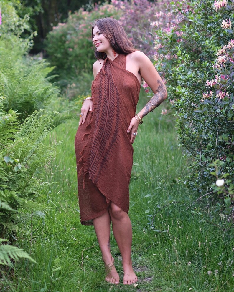 May include: A rust-coloured wrap dress featuring a halter neckline and a dark brown fern-like botanical print running vertically down the front. The garment has a textured fabric finish with fringe detailing along the side edge and a tie closure.