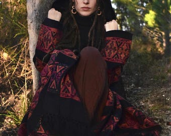 May include: A hooded black shawl with intricate red and orange geometric patterns. The shawl is draped over a black turtleneck and brown pants. The person is wearing gold hoop earrings and is sitting near a tree.