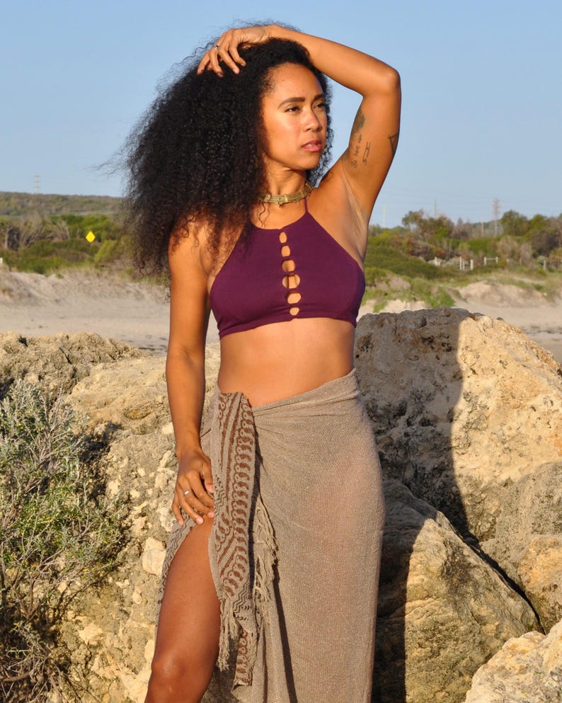 May include: A dark purple halter-style crop top featuring a vertical row of circular cutouts down the centre chest, paired with a semi-sheer, light brown sarong wrap that has a dark brown patterned border and fringe detailing along the side.