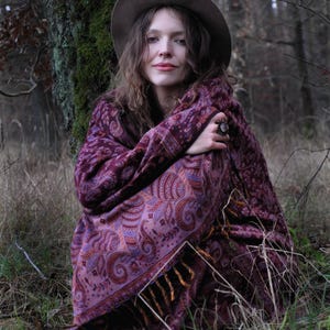 Könnte beinhalten: Eine Frau mit braunem Hut und bordeauxfarbenem Schal mit Paisley-Muster sitzt in einem Wald. Der Schal hat einen Fransen und die Farben umfassen Schattierungen von Lila, Orange und Braun. Die Frau sitzt in der Nähe eines Baumes.