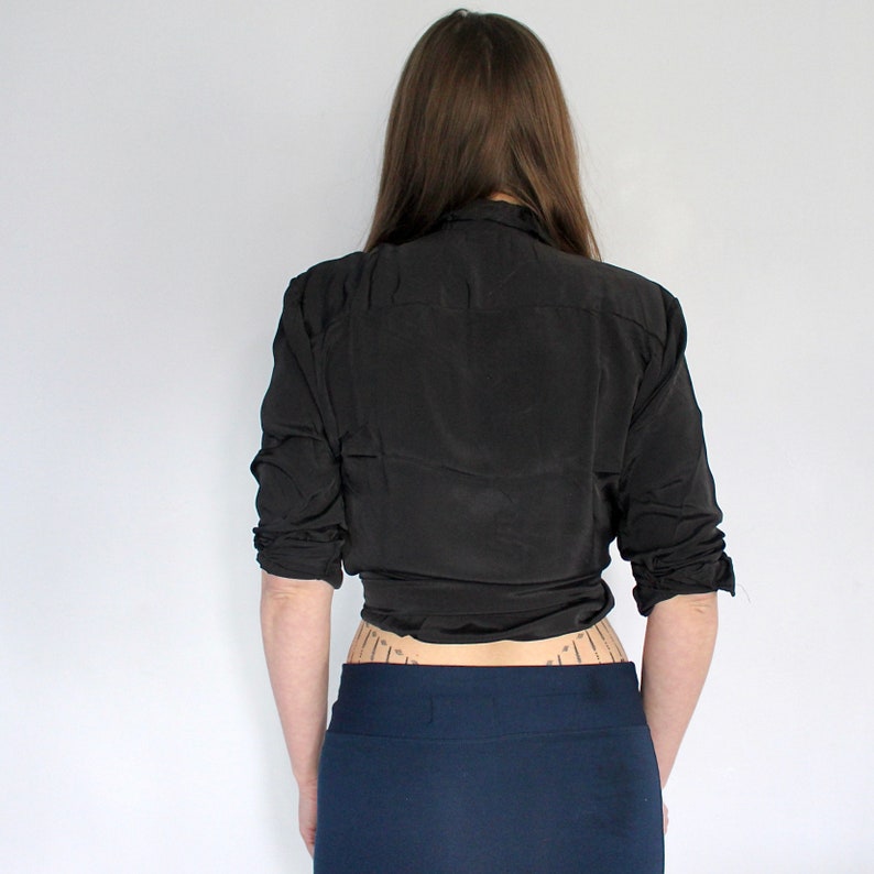 Puede incluir: Una camisa negra de manga larga con un frente de estilo envolvente. La camisa se usa sobre una falda azul oscuro.