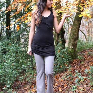 May include: A woman wearing a black tank top with a drawstring waist and gray pants. She is standing in a forest with fall leaves on the ground.