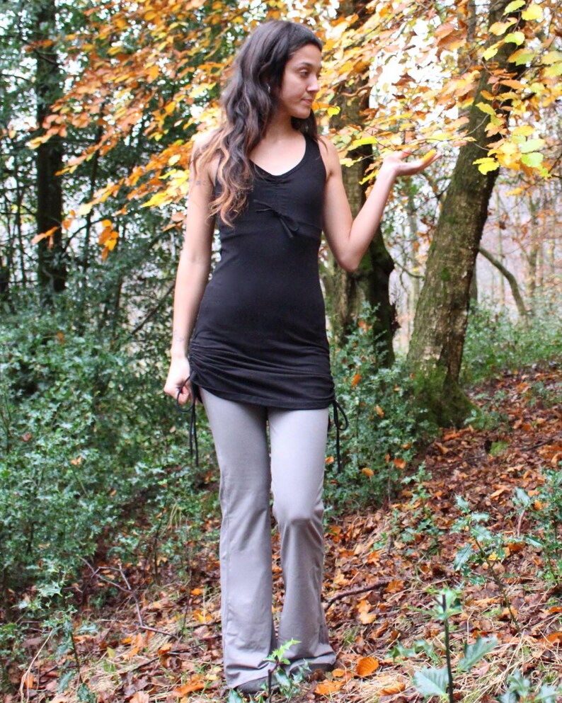 May include: A woman wearing a black tank top with a drawstring waist and gray pants. She is standing in a forest with fall leaves on the ground.
