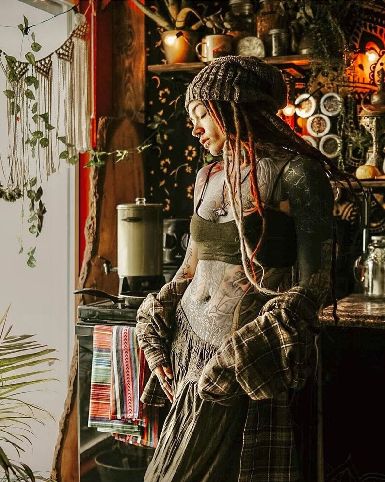 May include: A woman with long dreadlocks and tattoos is wearing a brown beanie, a green tank top, and a plaid shirt. She is standing in a kitchen with a wooden countertop and a variety of plants and decorations.