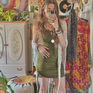 May include: A woman wearing a green, fitted, mini dress with fringe detailing. She is wearing a white crocheted cardigan and several bracelets on her wrist. She is standing in a room with a white cabinet and a patterned rug.