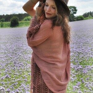 May include: A lightweight, dusty rose-coloured open-front cardigan featuring a sheer, loose-knit texture and a dark brown patterned border along the hem and sleeves. The garment is paired with a tan fedora hat featuring a dark, patterned band.