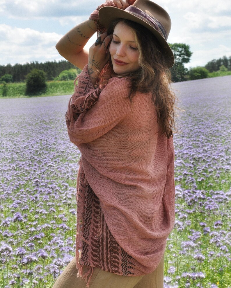 May include: A lightweight, dusty rose-coloured open-front cardigan featuring a sheer, loose-knit texture and a dark brown patterned border along the hem and sleeves. The garment is paired with a tan fedora hat featuring a dark, patterned band.