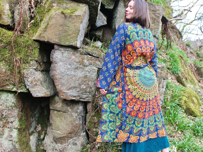 May include: A long, blue and orange patterned robe with a mandala design. The robe has a tie belt and long sleeves. The robe is worn by a person standing in front of a stone wall.